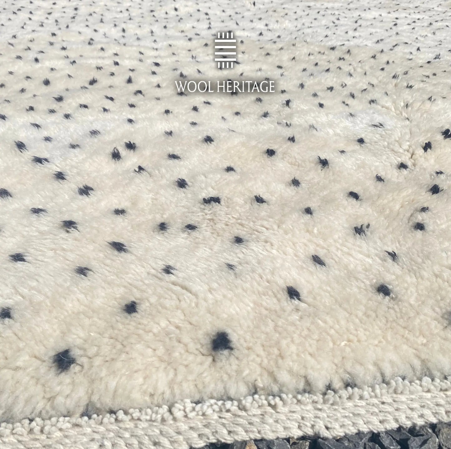Close-up of AMUD rug showing its soft ivory Moroccan wool pile, charcoal seed-like marks, and finished edge