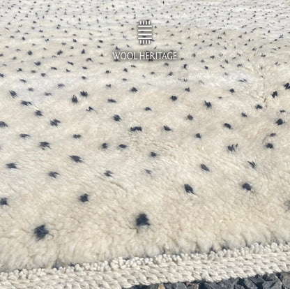 Close-up of AMUD rug showing its soft ivory Moroccan wool pile, charcoal seed-like marks, and finished edge