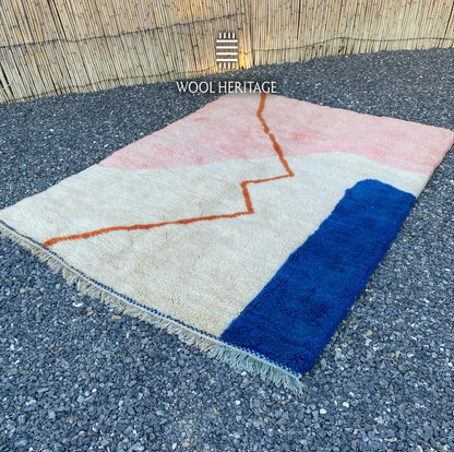 Angled view of TAYRI Moroccan rug showing its flowing terracotta line, ivory field, pink section, and blue side block