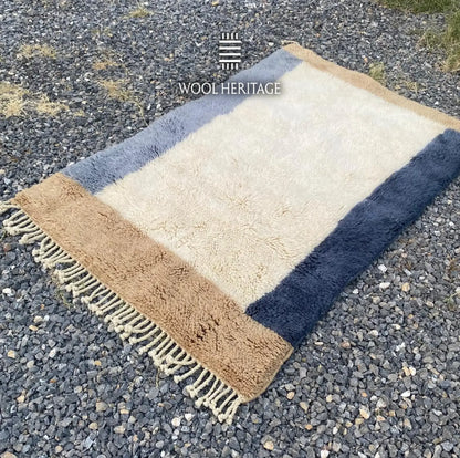 Angled view of TIFAWT Moroccan rug showing its luminous ivory field, blue side bands, beige ends, and fringe